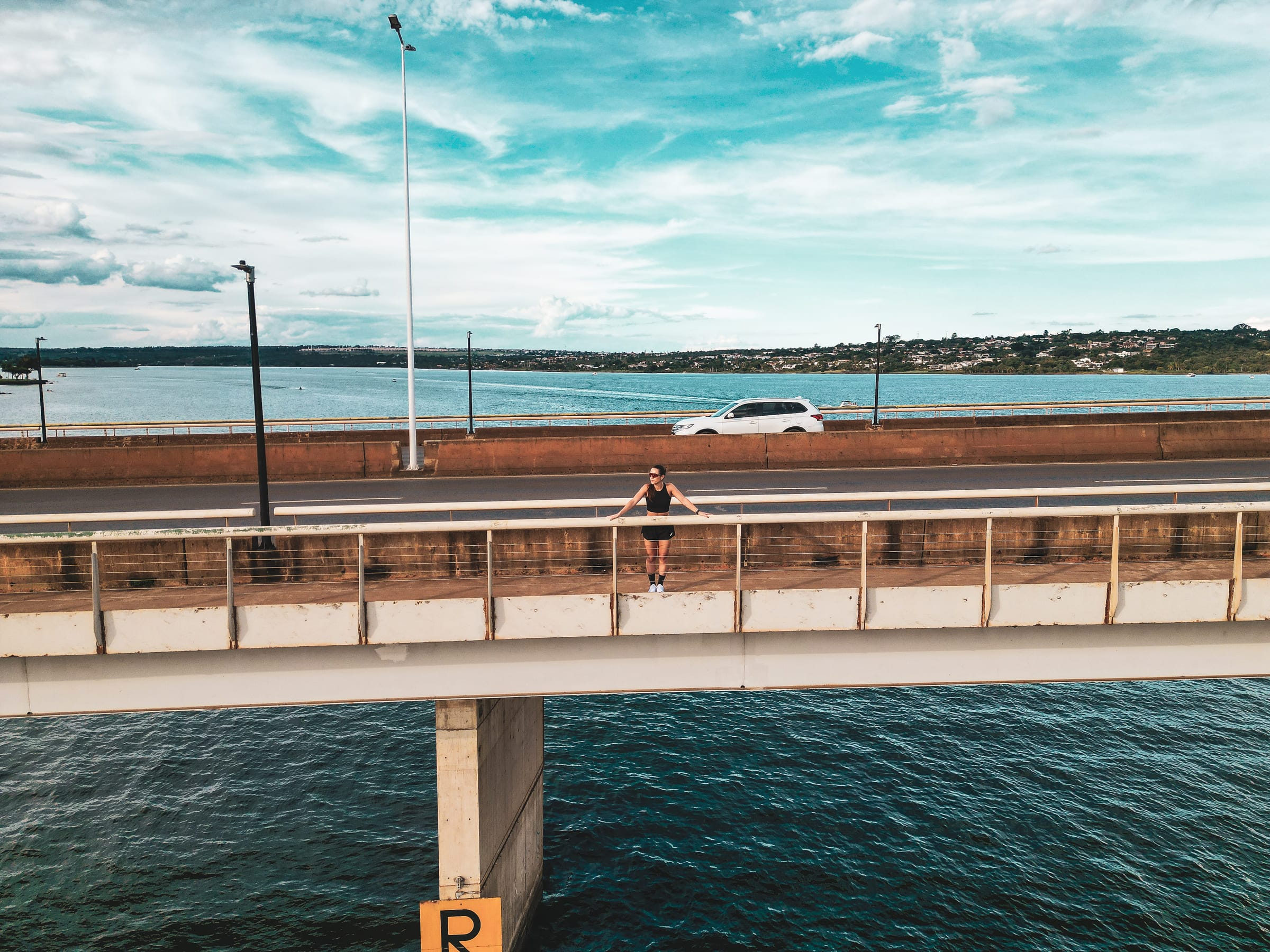 Vista aérea ampla da ponte sobre o Lago Paranoá, com Cristina apoiada no guarda-corpo