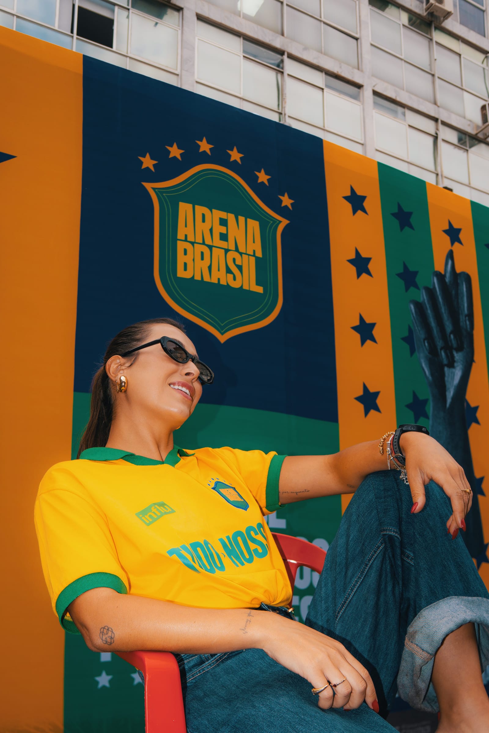 Cristina com a camisa amarela da seleção brasileira sentada diante de um mural azul e amarelo da Arena Brasil