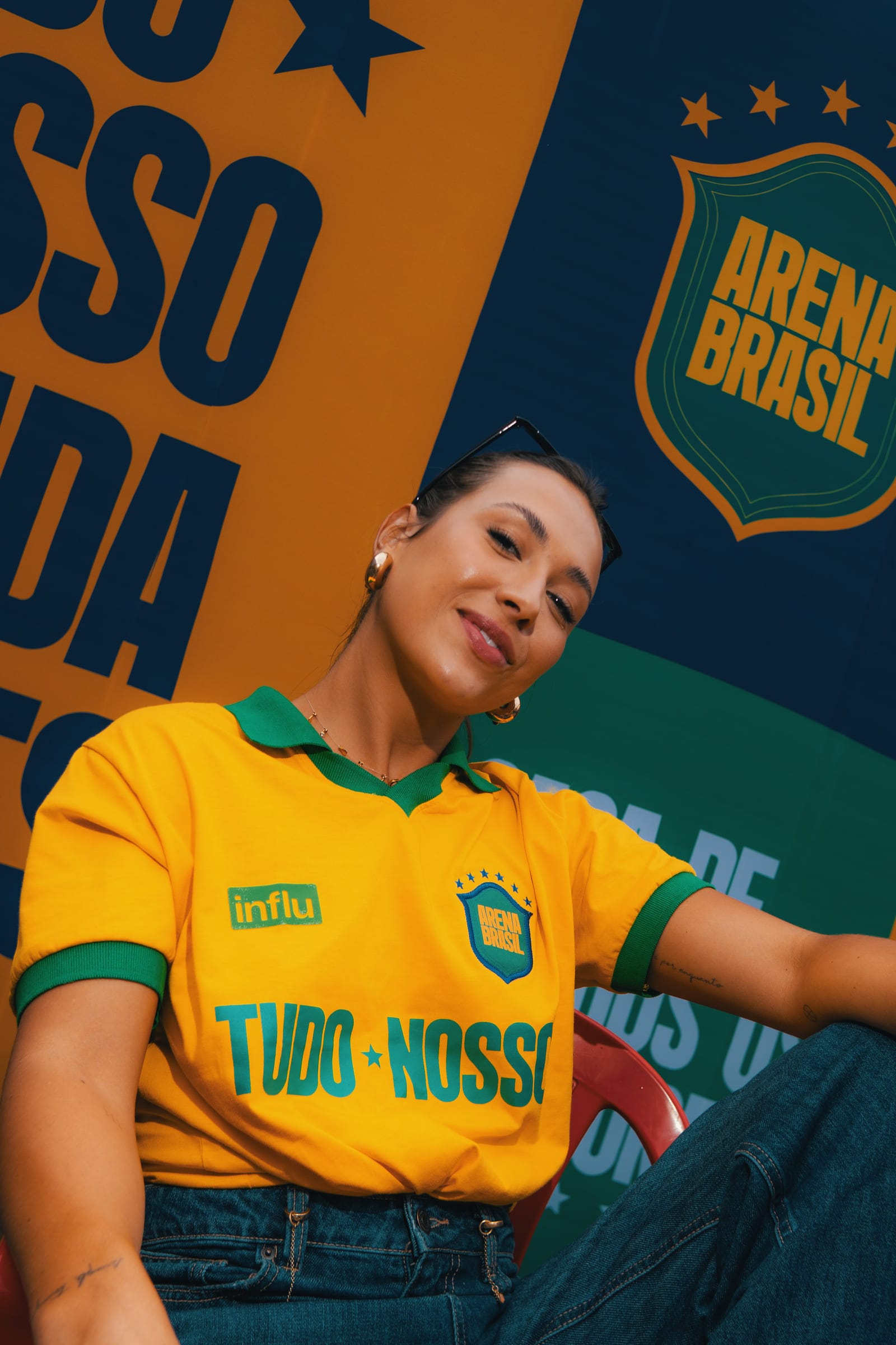 Retrato de Cristina de camisa da seleção brasileira, olhando para a câmera, com painéis coloridos ao fundo