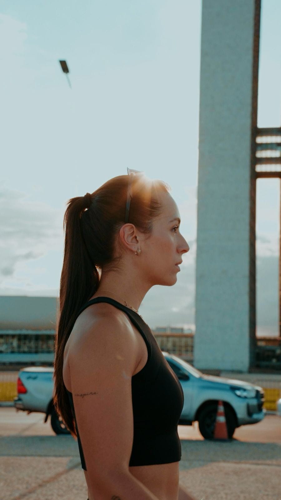Perfil de Cristina de rabo de cavalo e top preto, em contraluz de fim de tarde em Brasília