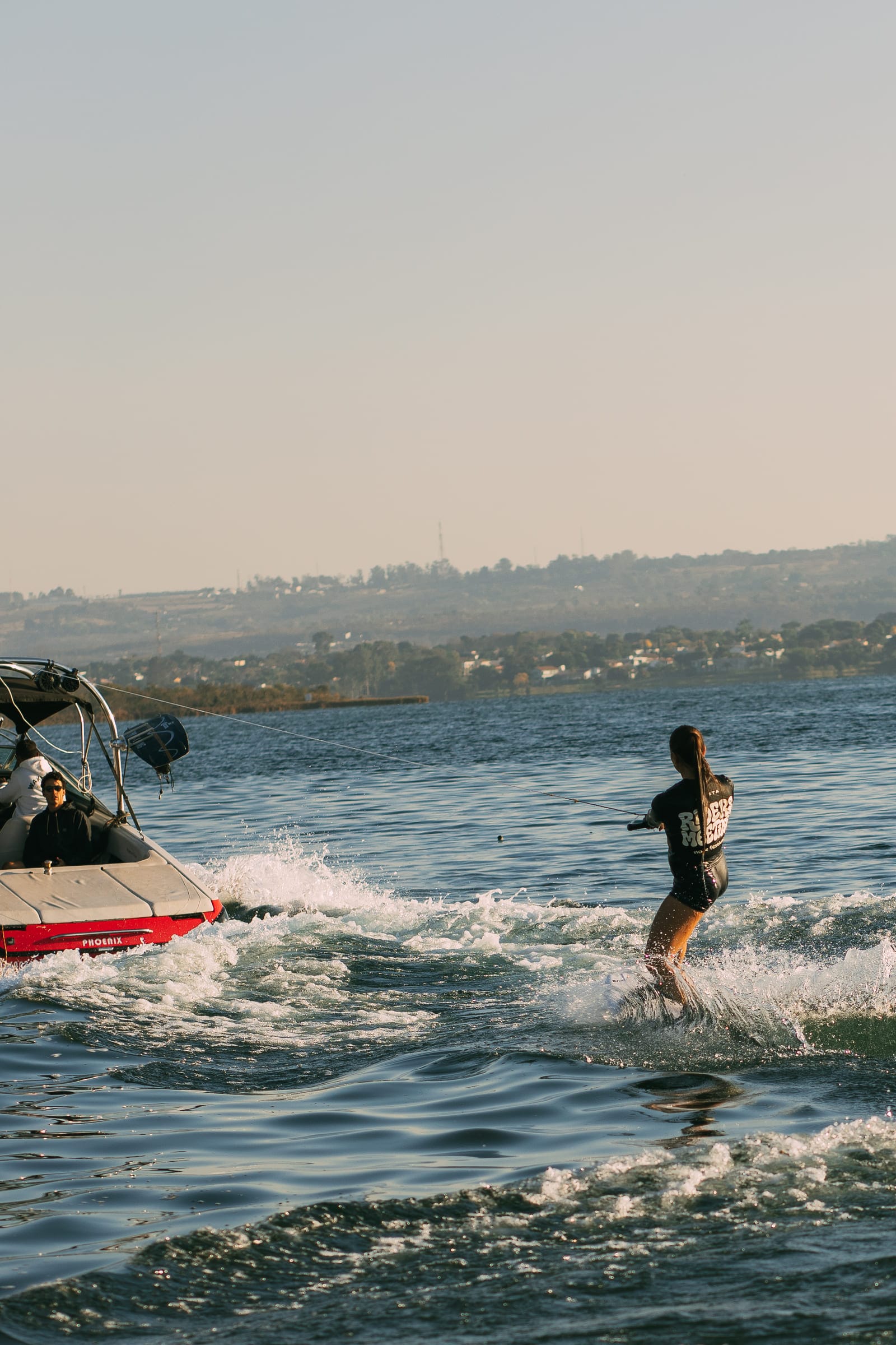 Cristina de wakeboard atrás do barco, em silhueta sobre a água dourada do fim de tarde